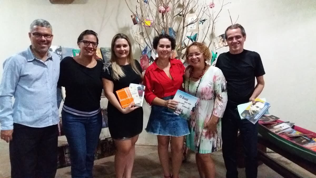 Seis pessoas sorriem segurando livros em frente a uma árvore decorada e a uma estante de livros