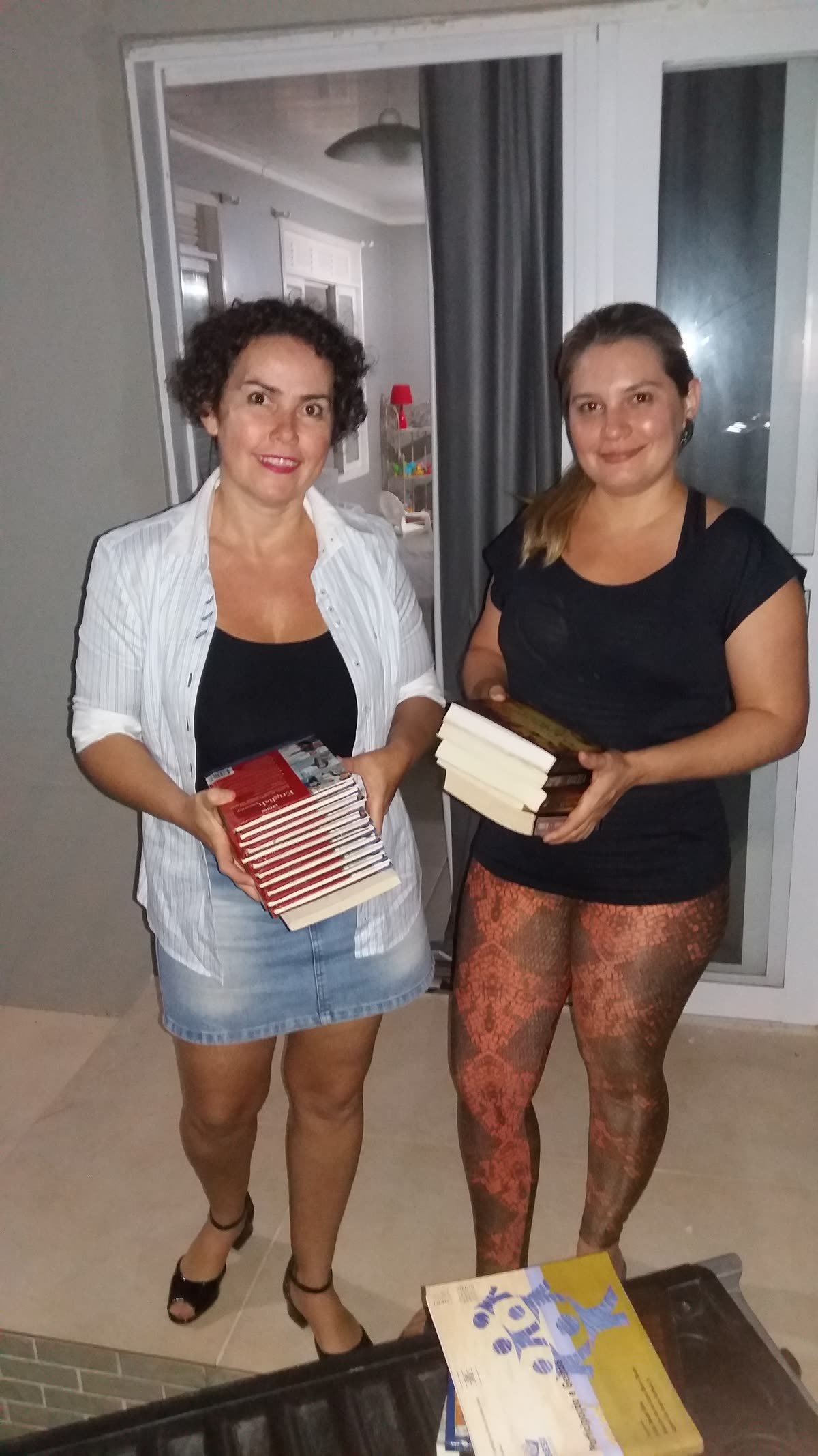 Duas mulheres seguram pilhas de livros na porta de uma casa
