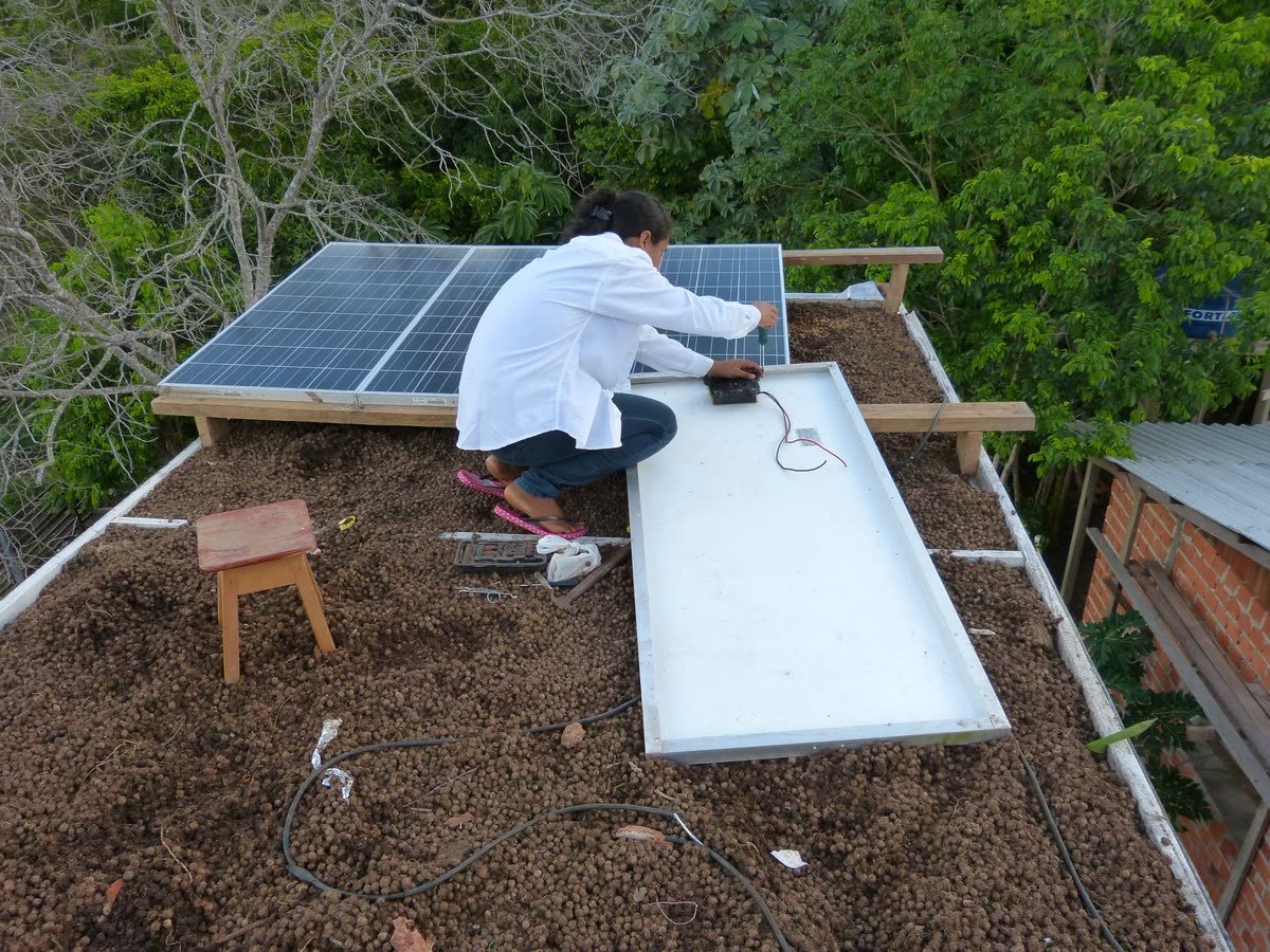 Pessoa trabalha sobre o telhado coberto de substrato, junto a placas solares, cercado de árvores