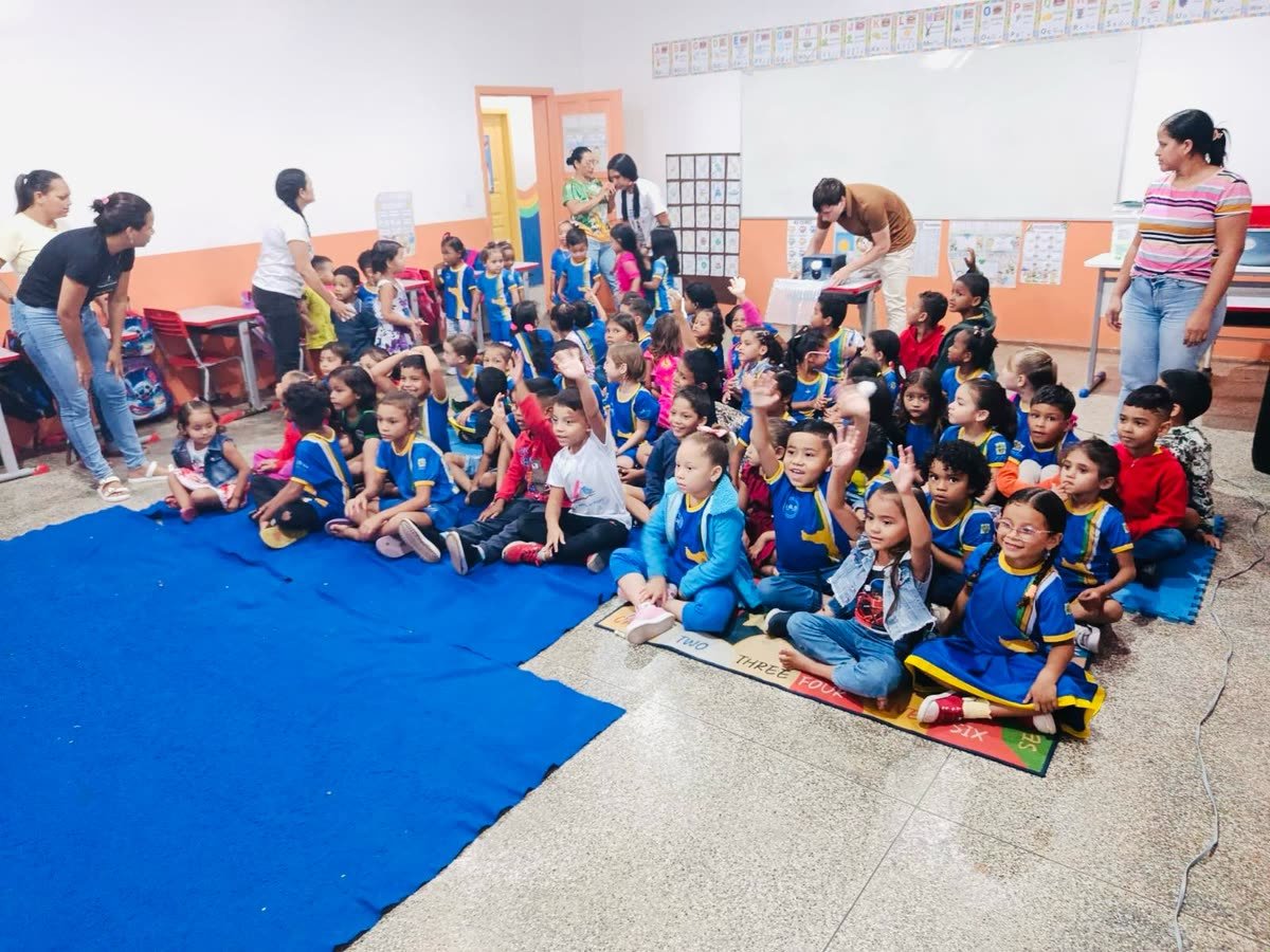 Turma de crianças pequenas sentadas no chão de uma sala de aula, algumas com a mão levantada, antes da sessão