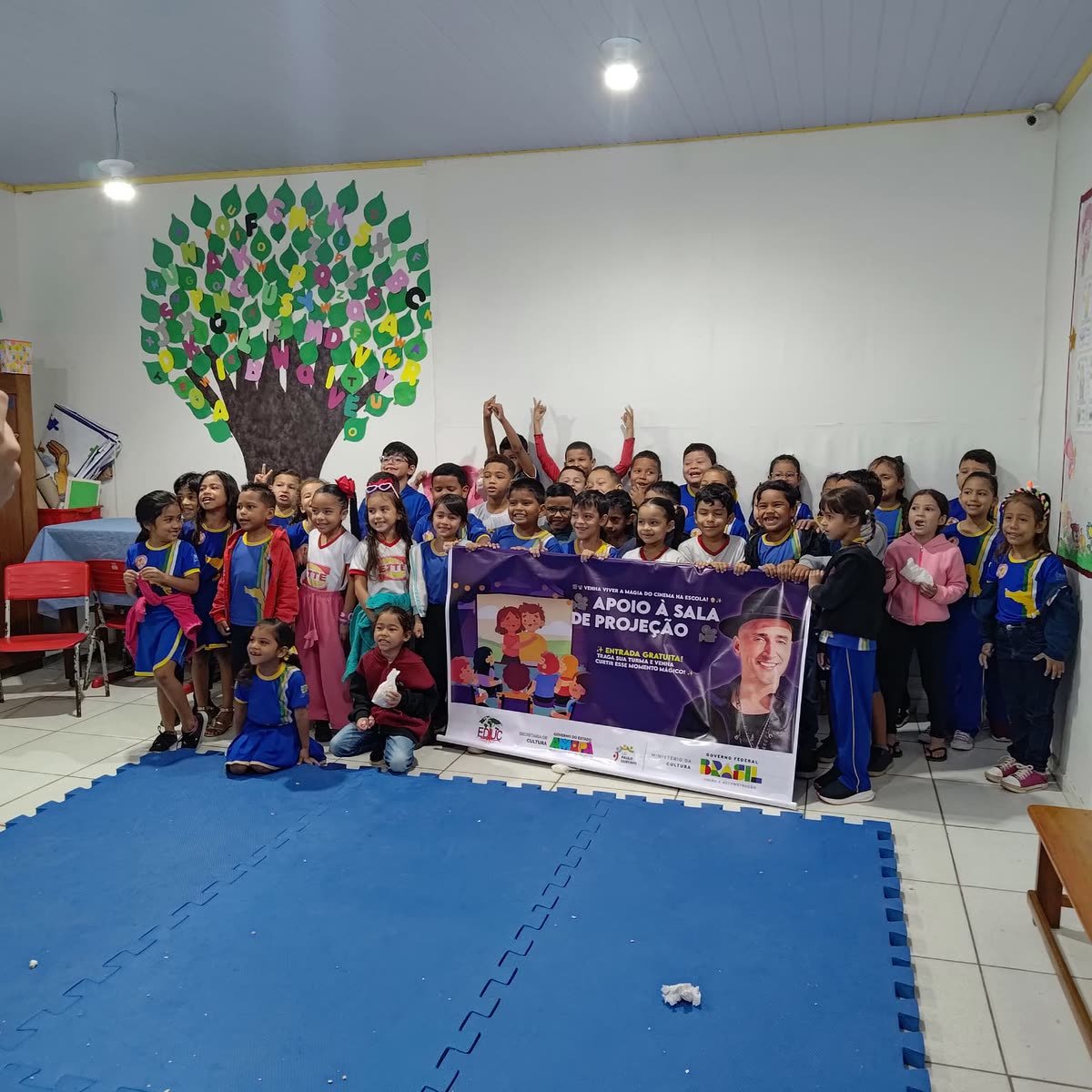 Crianças posam atrás de uma faixa da sala de projeção em uma sala decorada com uma árvore de letras