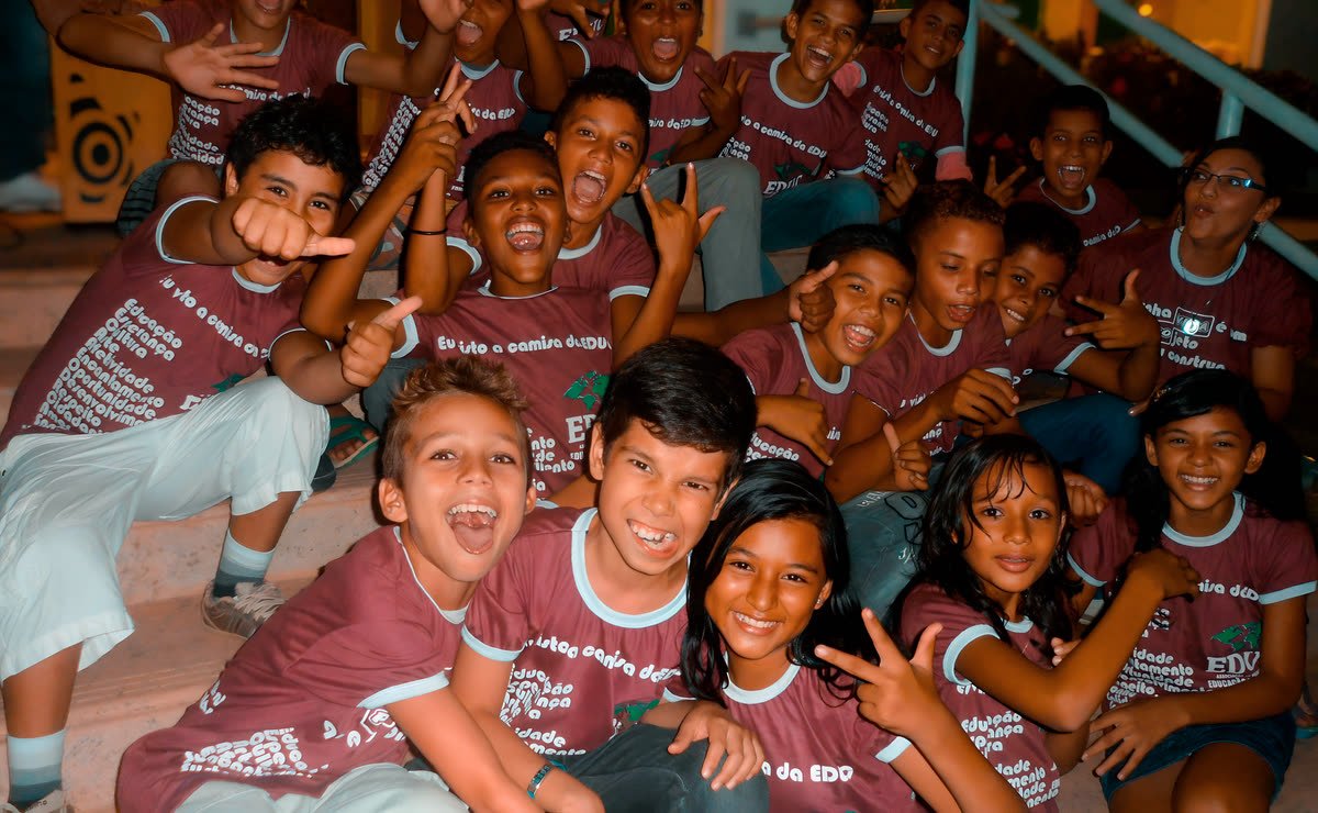 Grupo de crianças e adolescentes com camisetas da EDUC sorri e faz gestos para a câmera em uma escadaria