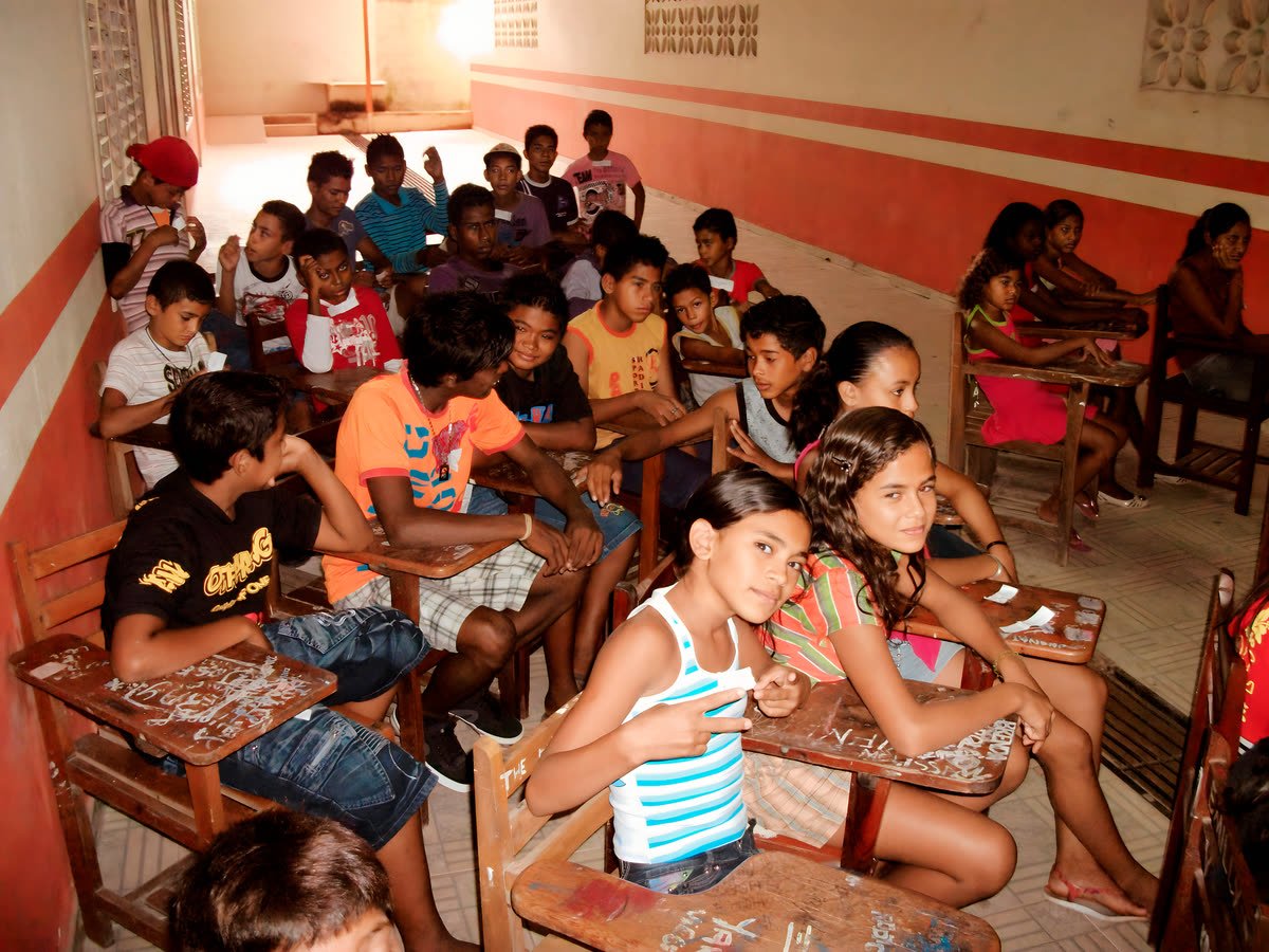 Crianças e adolescentes sentados em carteiras escolares em um corredor de escola