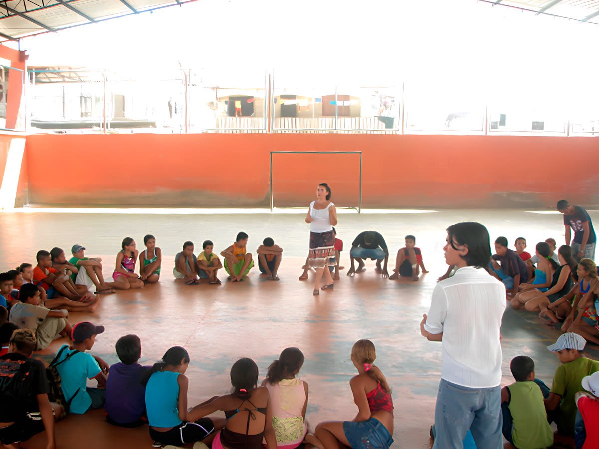Crianças e adolescentes sentados em roda no chão de uma quadra, ouvindo uma educadora