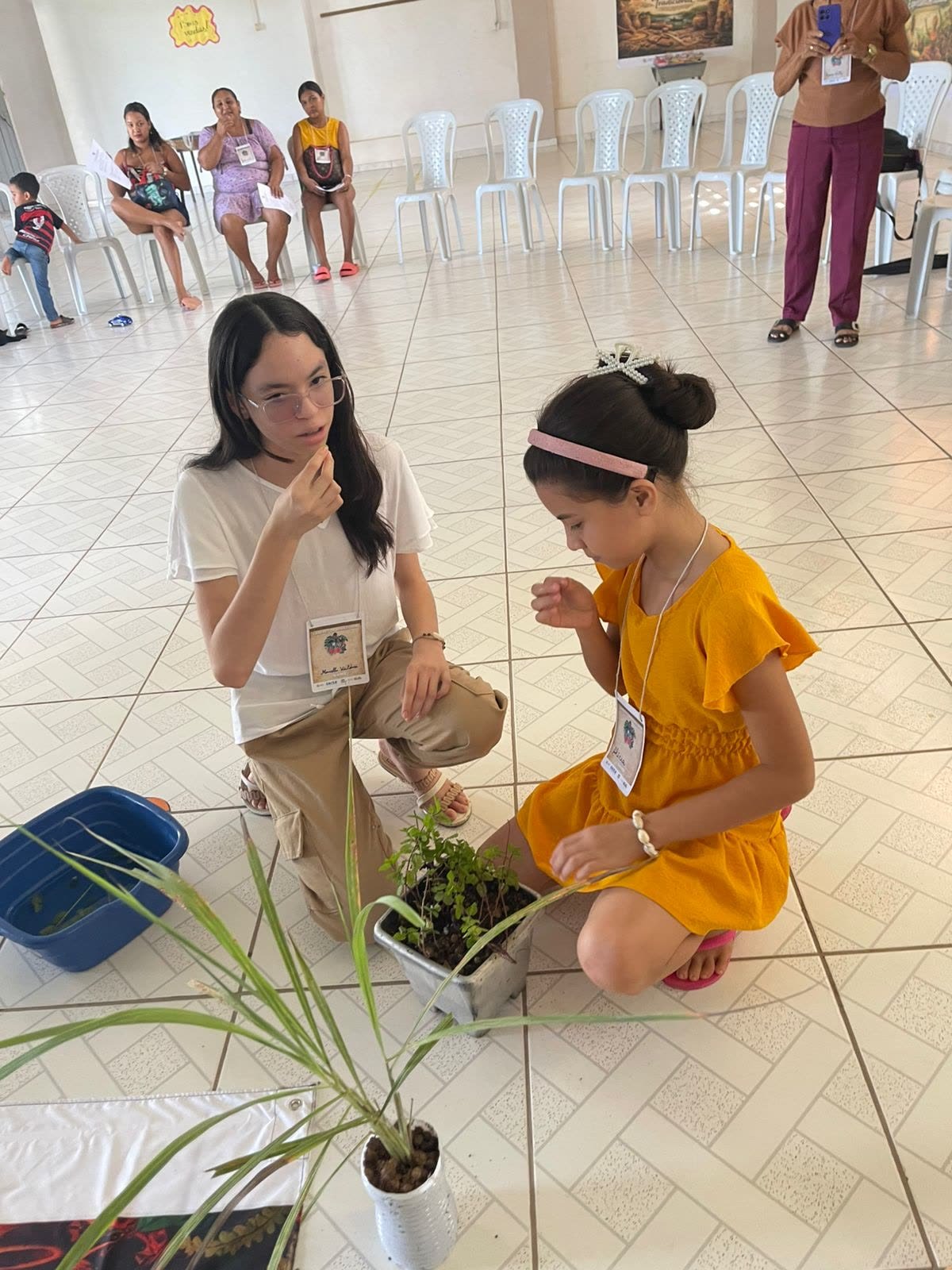 Duas participantes ajoelhadas junto a uma planta em vaso durante a atividade