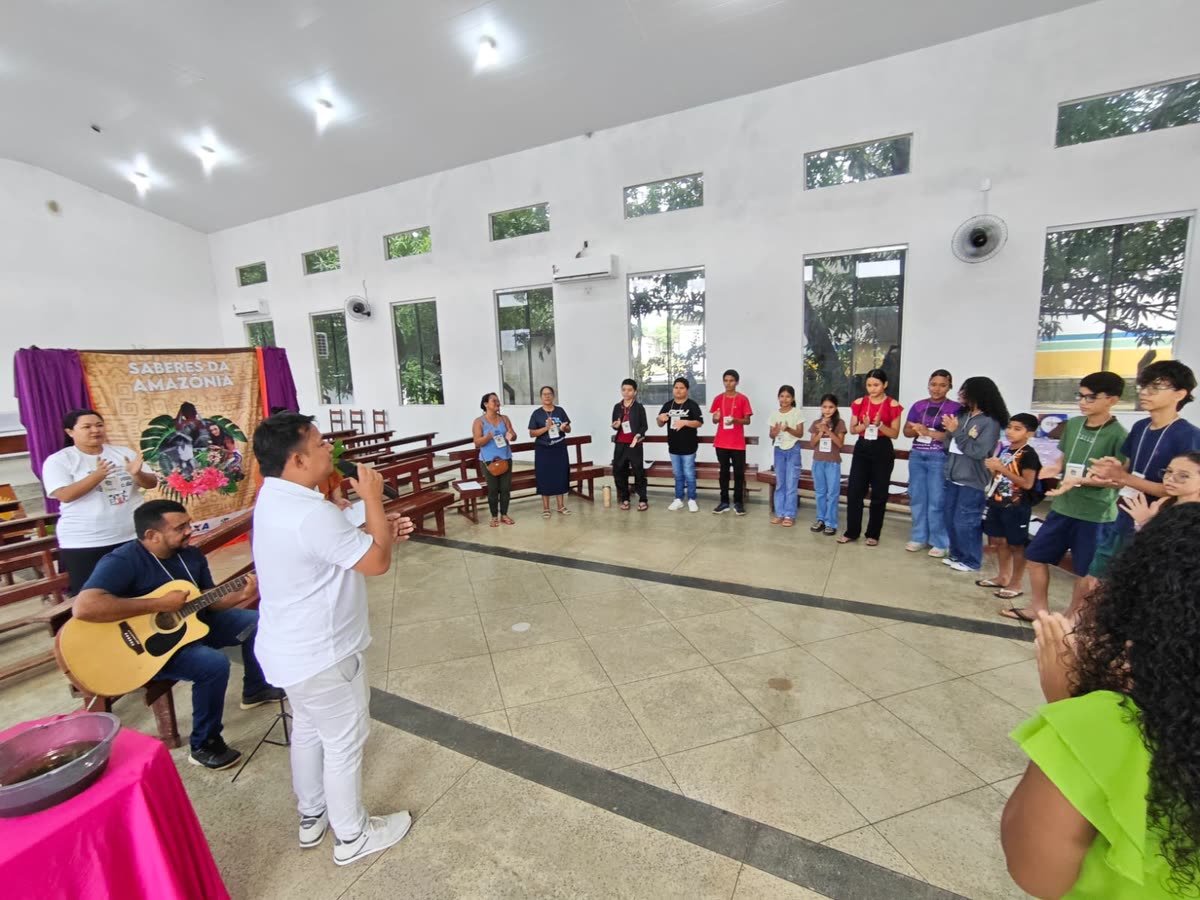 Participantes em roda batem palmas acompanhados por violão, ao lado da faixa Saberes da Amazônia