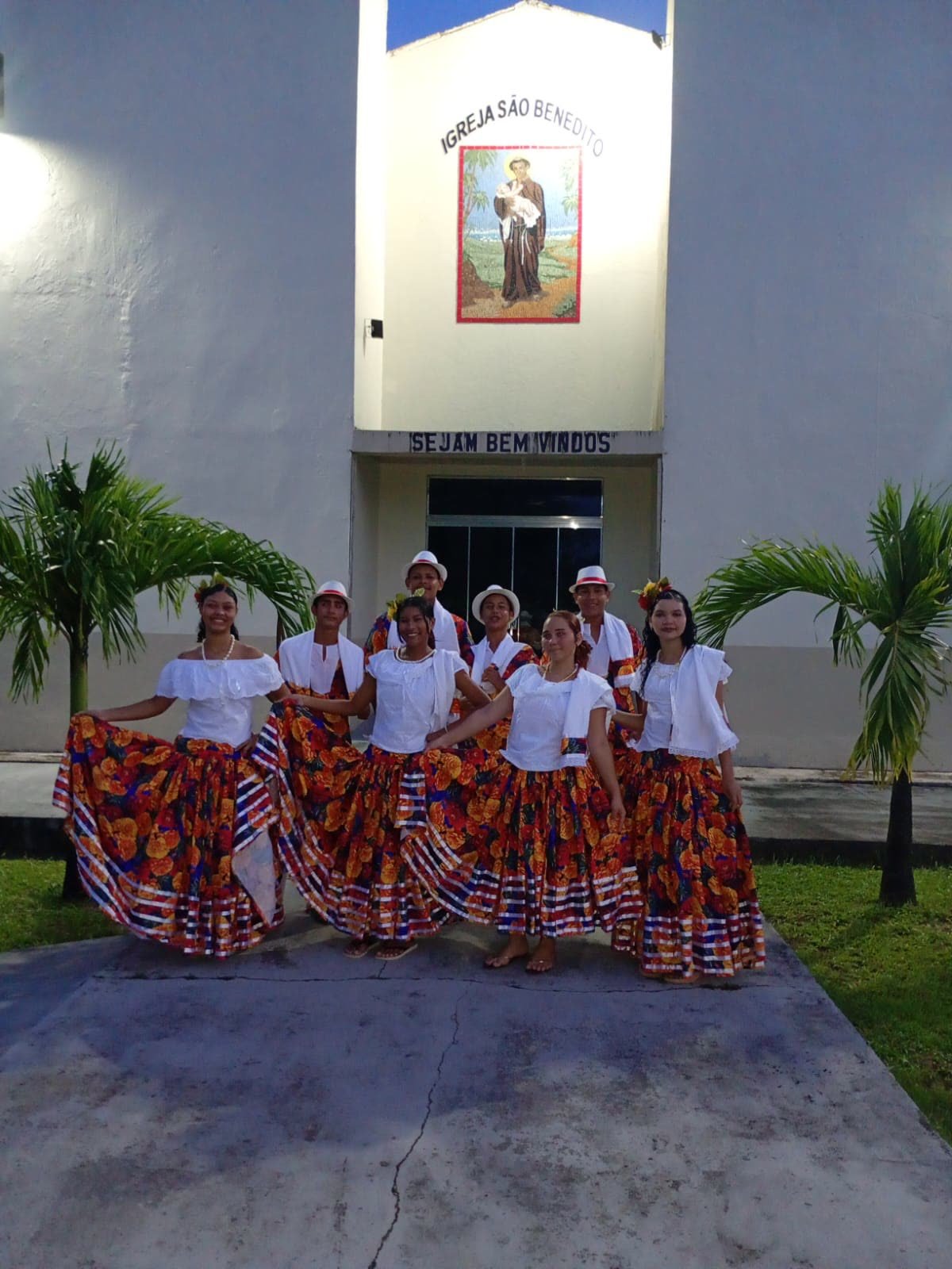 Grupo de dançarinos com saias floridas e chapéus brancos em frente a uma igreja